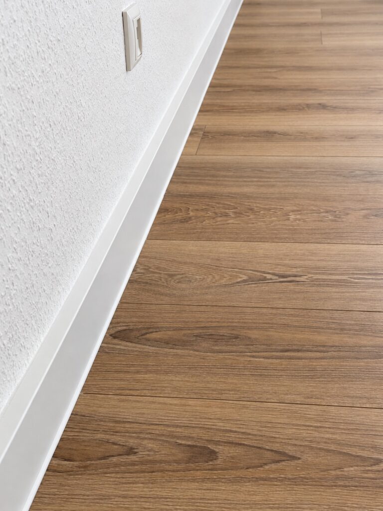 Weiße Sockelleiste entlang einer Wand mit Steckdose und Boden in Holzoptik