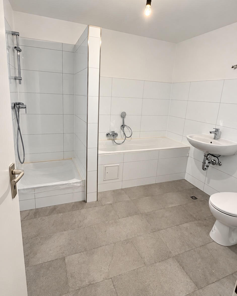 Bad mit weißen Wandfliesen, grauem Boden, Dusche und Badewanne; Schutzfolie an der Dusche und provisorische Beleuchtung.