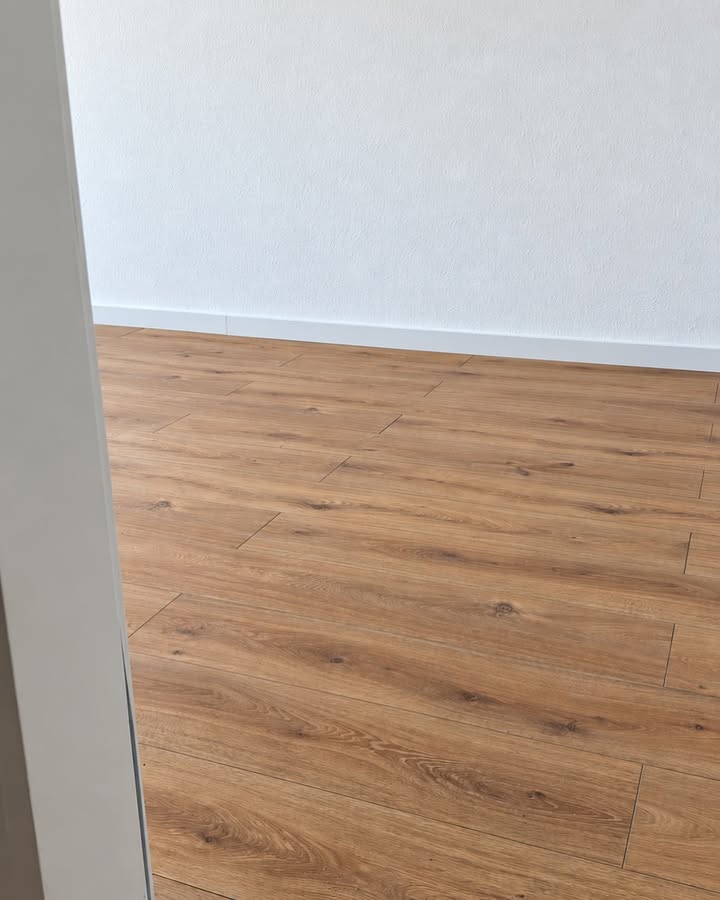 Boden in Holzoptik mit weißen Sockelleisten und heller Wand, durch eine Türöffnung fotografiert.